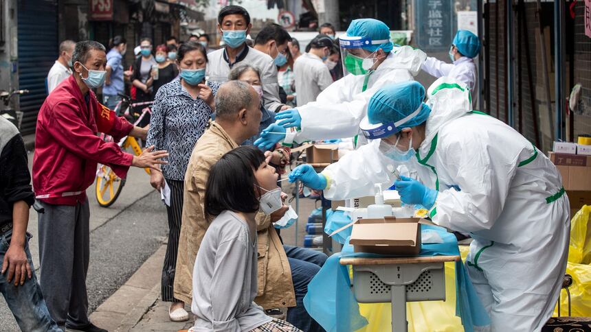 Health workers in full protective equipment swab seated residents at a street-side booth in Wuhan, with a crowd of masked people waiting behind them.